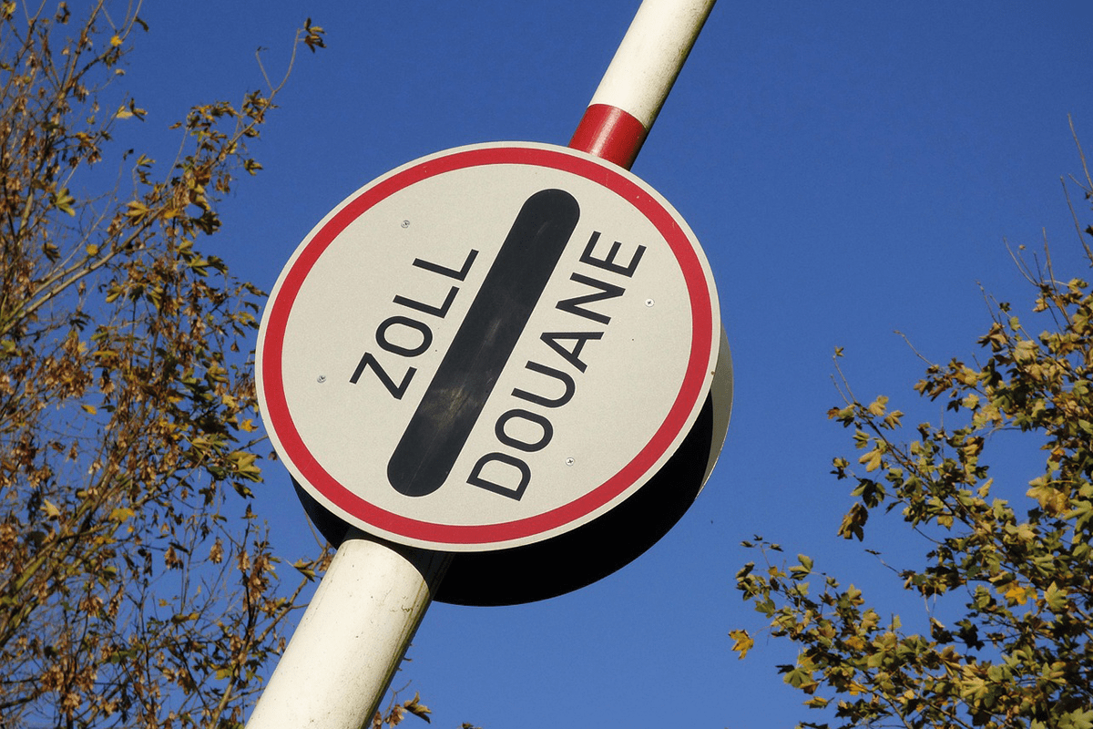 Rundes Verkehrszeichen mit der Aufschrift „Zoll/Douane“ vor blauem Himmel und herbstlichen Bäumen.