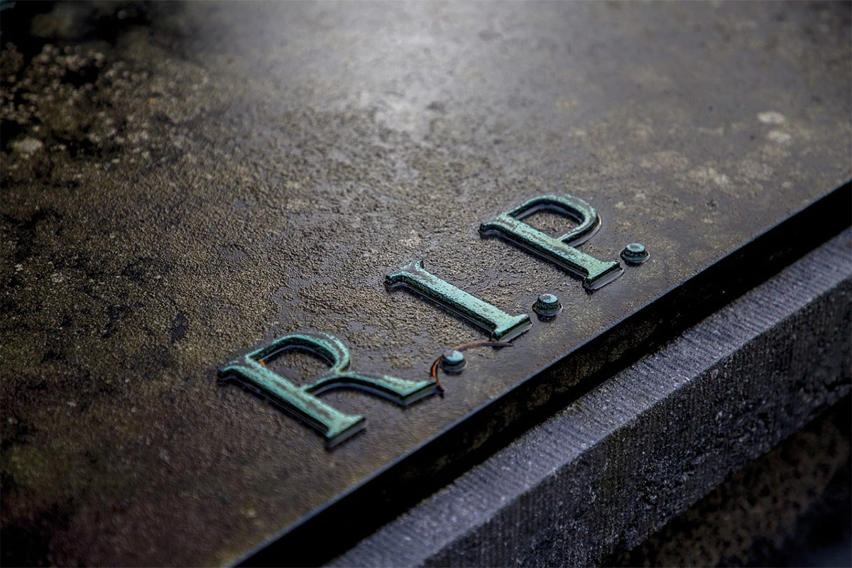 Nahaufnahme eines Grabsteins mit erhabener, grüner R.I.P.-Inschrift auf dunklem Stein.