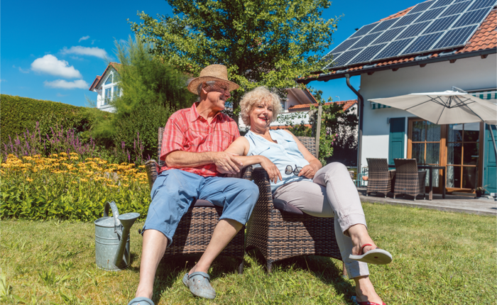 Seniorenpaar sitzt entspannt im Garten vor einem Haus mit Solaranlage bei sonnigem Wetter.