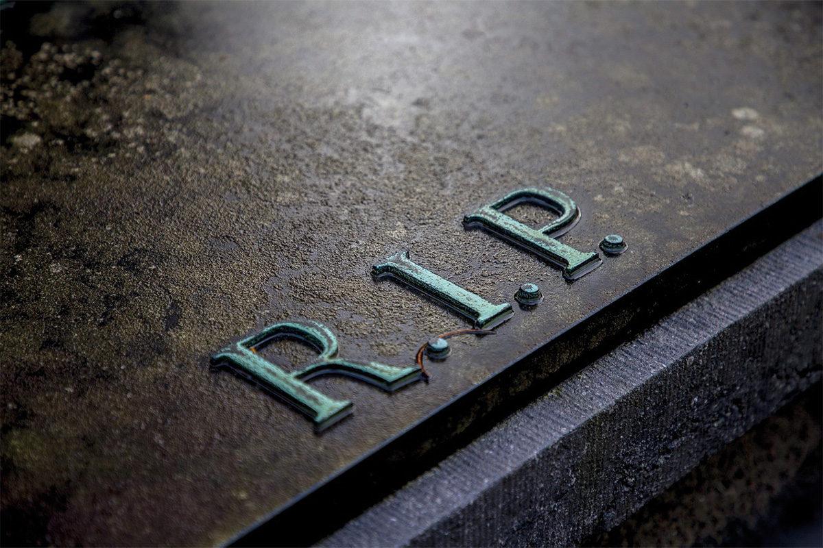 Nahaufnahme eines Grabsteins mit erhabener, grüner R.I.P.-Inschrift auf dunklem Stein.