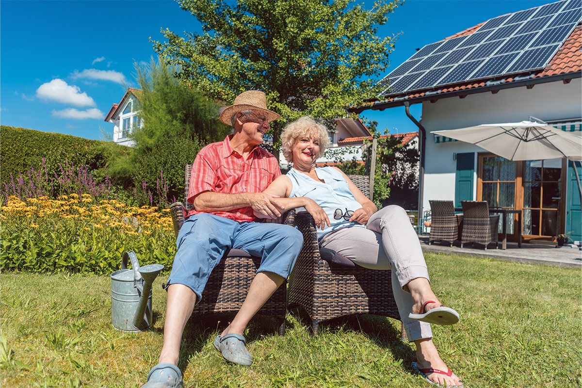 Seniorenpaar sitzt entspannt im Garten vor einem Haus mit Solaranlage bei sonnigem Wetter.