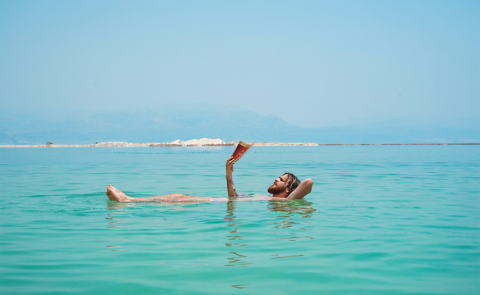 Mann liegt enspannt lesend im Meer mit Küste im Hintergrund.