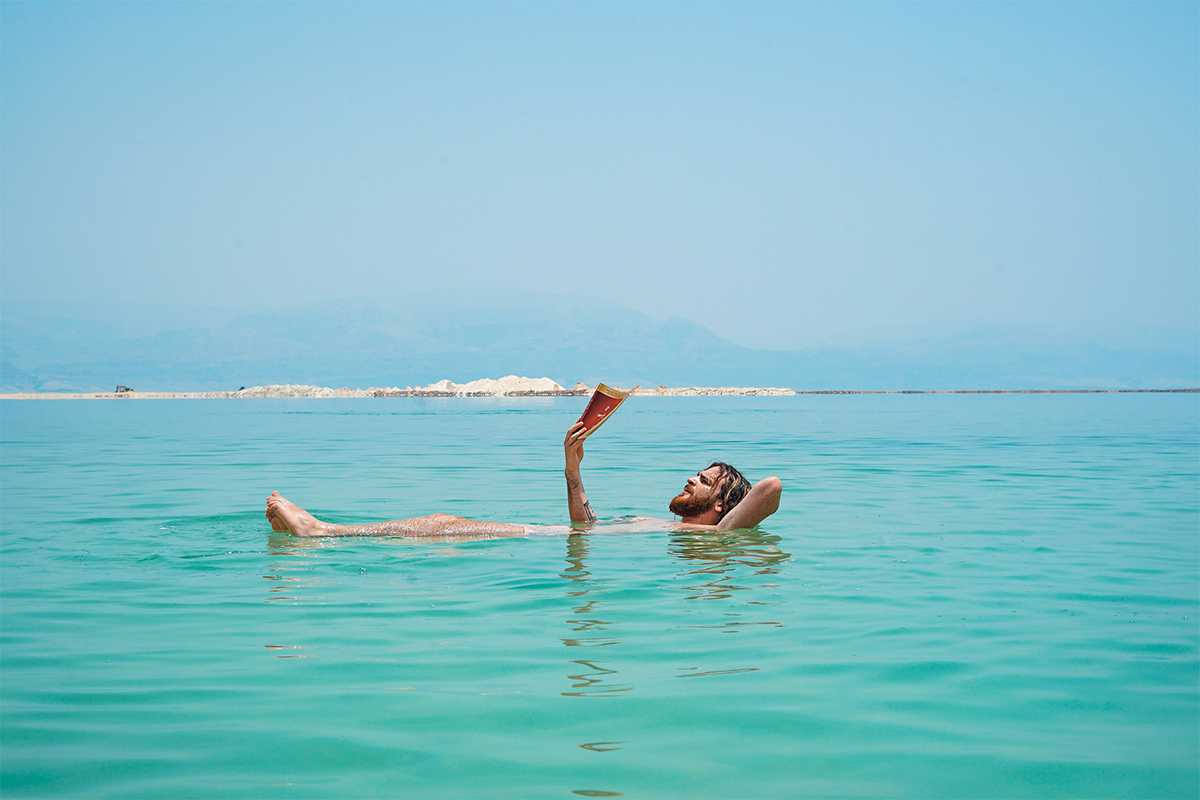 Mann liegt enspannt lesend im Meer mit Küste im Hintergrund.