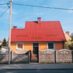 Einfamilienhaus mit rotem Ziegeldach und gelber Fassade hinter einem Steinzaun mit weißen Gittern.