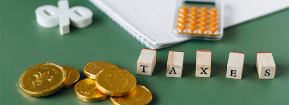 Goldmünzen, Holzwürfel mit dem Wort "TAXES", ein Block und ein Taschenrechner liegen auf einem grünen Tisch.